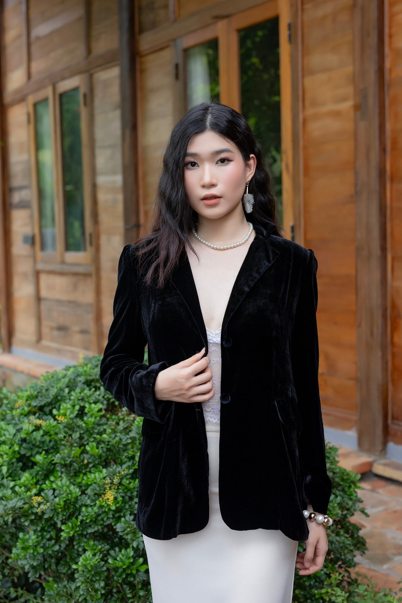 Pode incluir: Uma mulher vestindo um blazer de veludo preto com um colar e brincos de p&eacute;rolas. Ela est&aacute; em p&eacute; na frente de um pr&eacute;dio de madeira com uma saia branca.