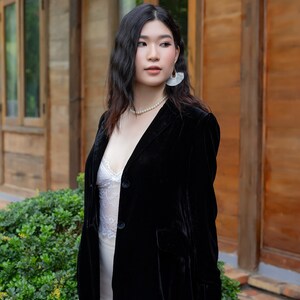 Pode incluir: Uma mulher vestindo um blazer de veludo preto sobre uma blusa de renda branca e uma saia branca. Ela est&aacute; usando brincos de p&eacute;rola e um colar de p&eacute;rola. Ela est&aacute; em p&eacute; na frente de uma porta de madeira.