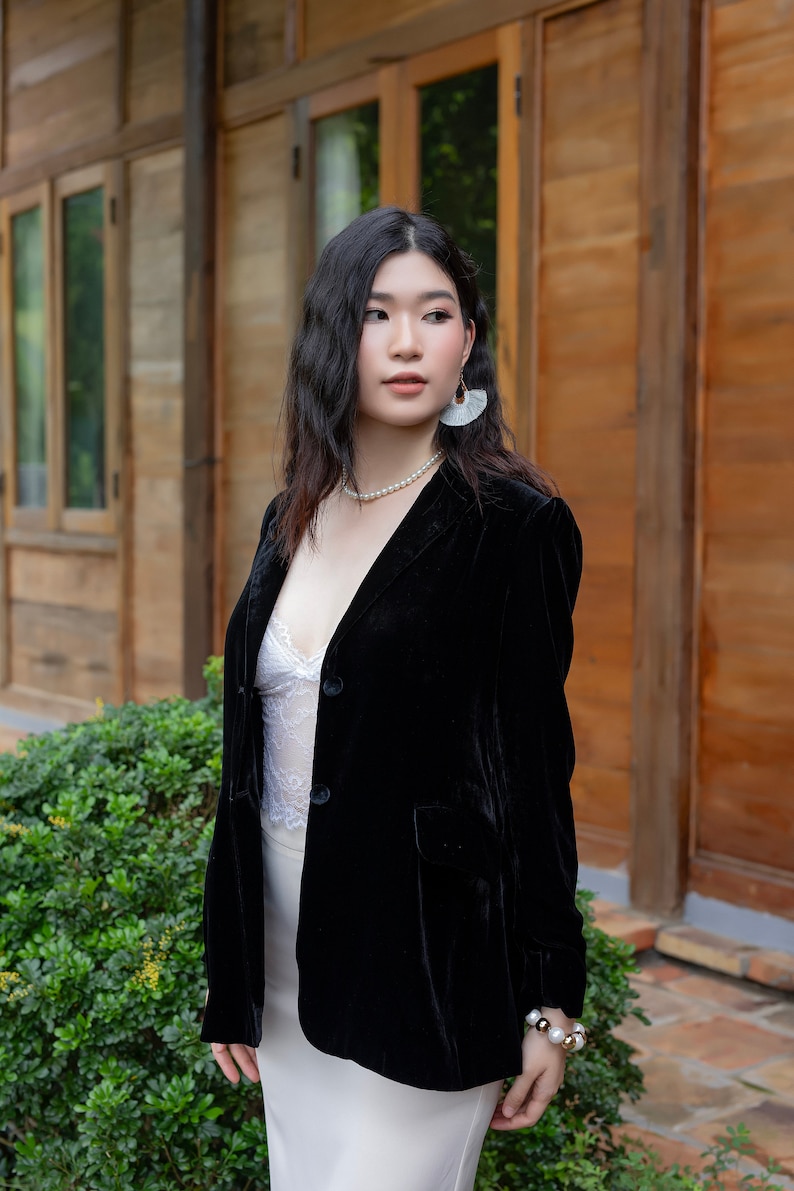 Pode incluir: Uma mulher vestindo um blazer de veludo preto sobre uma blusa de renda branca e uma saia branca. Ela est&aacute; usando brincos de p&eacute;rola e um colar de p&eacute;rola. Ela est&aacute; em p&eacute; na frente de uma porta de madeira.