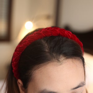 May include: A red velvet headband with a braided design. The headband is worn on a person's head.