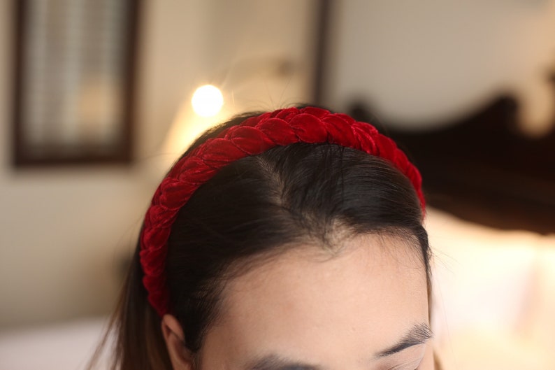 May include: A red velvet headband with a braided design. The headband is worn on a person's head.