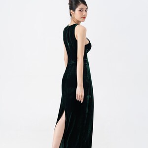 May include: A sleeveless, dark green velvet gown with a high slit. The dress has a fitted bodice and a flowing skirt. The model is wearing nude-colored high heels. The background is plain white.
