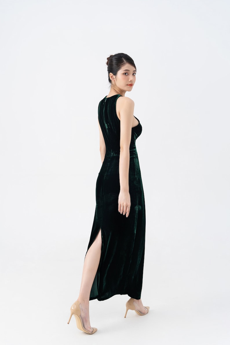 May include: A sleeveless, dark green velvet gown with a high slit. The dress has a fitted bodice and a flowing skirt. The model is wearing nude-colored high heels. The background is plain white.