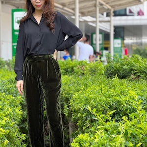 May include: A woman wearing a black button-down shirt and dark green velvet pants. She is standing in front of a green bush.