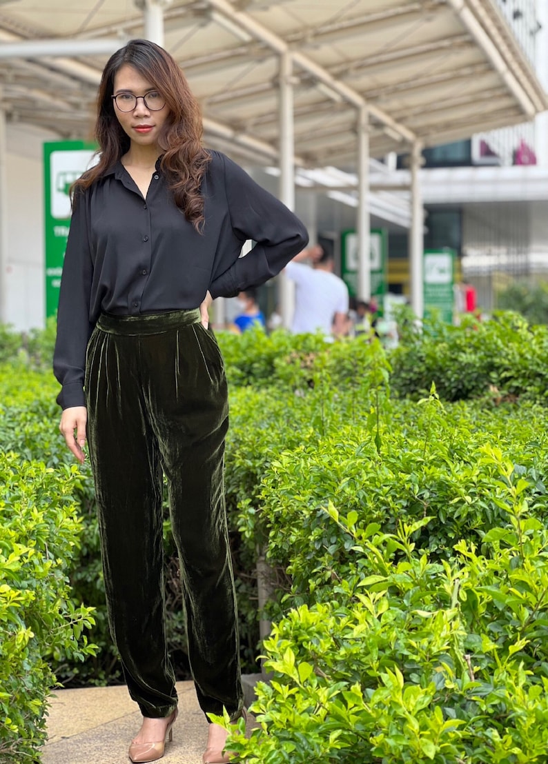 May include: A woman wearing a black button-down shirt and dark green velvet pants. She is standing in front of a green bush.