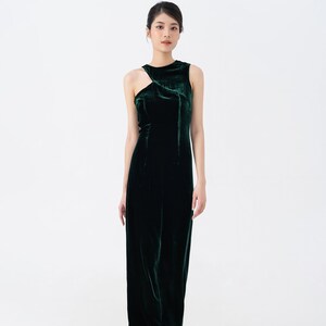 May include: A sleeveless, floor-length, dark green velvet dress. The dress features a unique asymmetrical neckline with a cutout detail on one shoulder. The fabric has a soft sheen, and the dress is form-fitting.