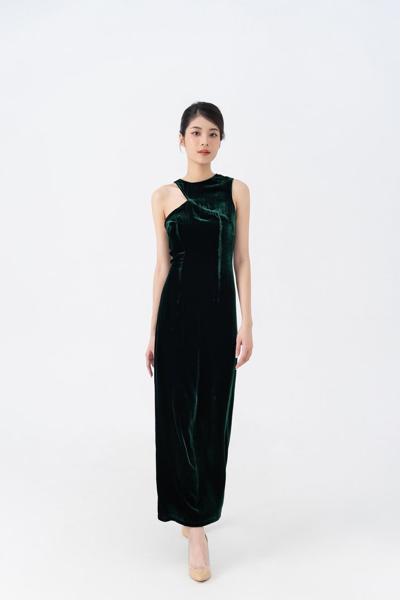 May include: A sleeveless, floor-length, dark green velvet dress. The dress features a unique asymmetrical neckline with a cutout detail on one shoulder. The fabric has a soft sheen, and the dress is form-fitting.