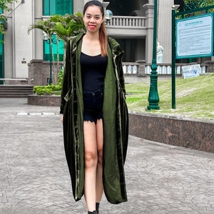 May include: A woman wearing a long, dark green velvet duster coat with a black tank top and denim shorts. She is wearing black ankle boots and has long dark hair.