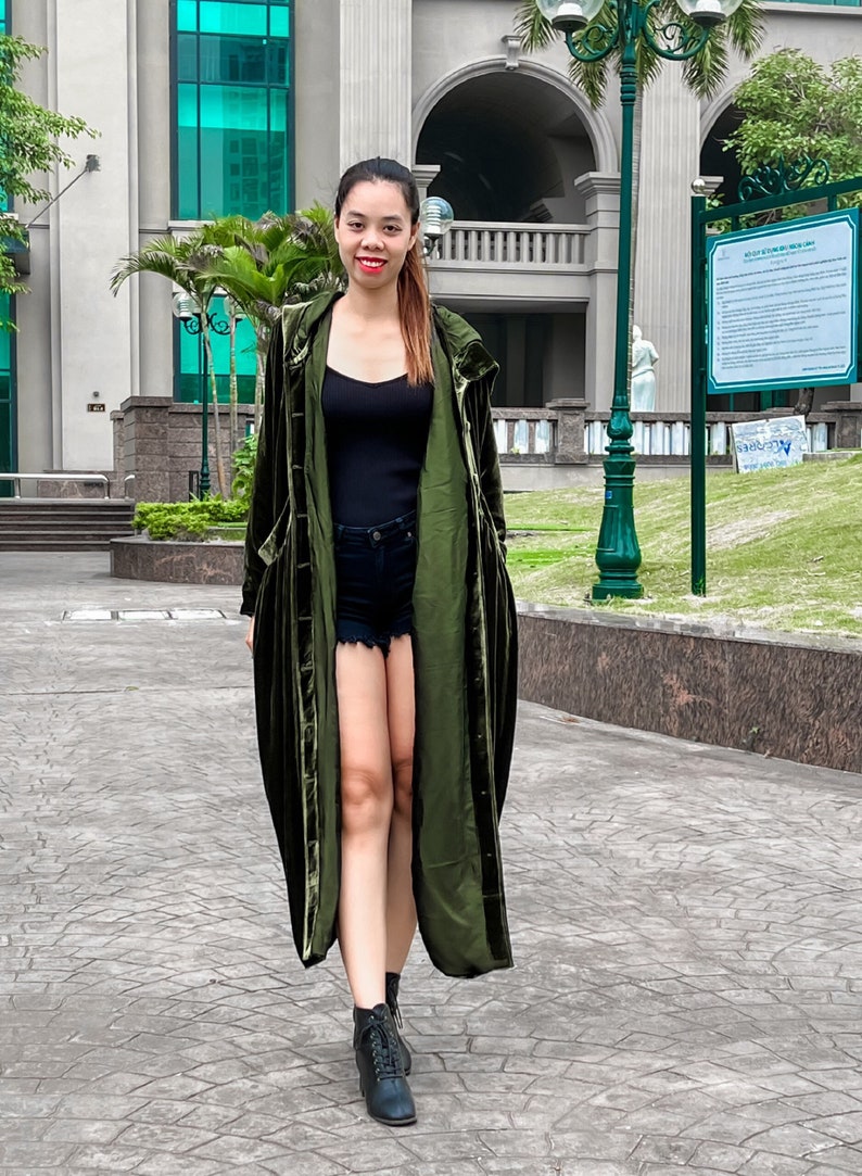 May include: A woman wearing a long, dark green velvet duster coat with a black tank top and denim shorts. She is wearing black ankle boots and has long dark hair.