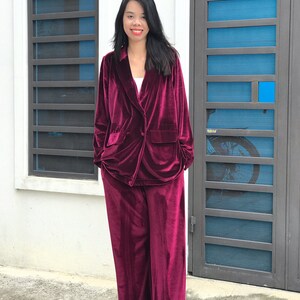 May include: A woman wearing a burgundy velvet blazer and matching pants. The blazer has a double-breasted closure and a pocket on the left side. The pants are wide-legged and have a straight fit.
