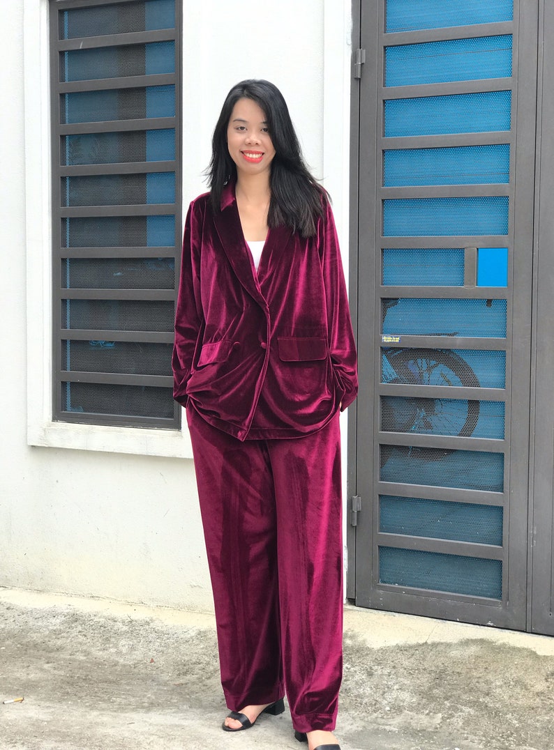 May include: A woman wearing a burgundy velvet blazer and matching pants. The blazer has a double-breasted closure and a pocket on the left side. The pants are wide-legged and have a straight fit.