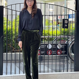 May include: A woman wearing a black collared shirt and dark green velvet pants. She is standing in front of a black metal gate.