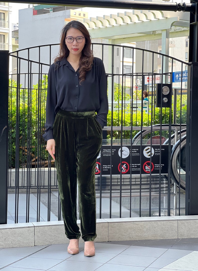 May include: A woman wearing a black collared shirt and dark green velvet pants. She is standing in front of a black metal gate.