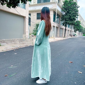 May include: A long, light green velvet dress with a long sleeve and a slightly puffed sleeve. The dress has a V-neckline and a flowy skirt. The model is wearing white sneakers.