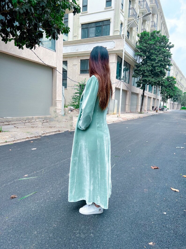 May include: A long, light green velvet dress with a long sleeve and a slightly puffed sleeve. The dress has a V-neckline and a flowy skirt. The model is wearing white sneakers.
