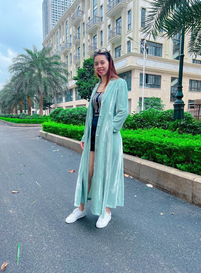May include: A woman wearing a long, light green velvet coat with a black and white checkered top and black shorts. She is standing on a paved walkway in front of a building.