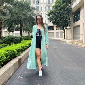 May include: A woman wearing a long, light green velvet duster coat over a black and white checkered top and black shorts. She is walking on a paved path with a building in the background.