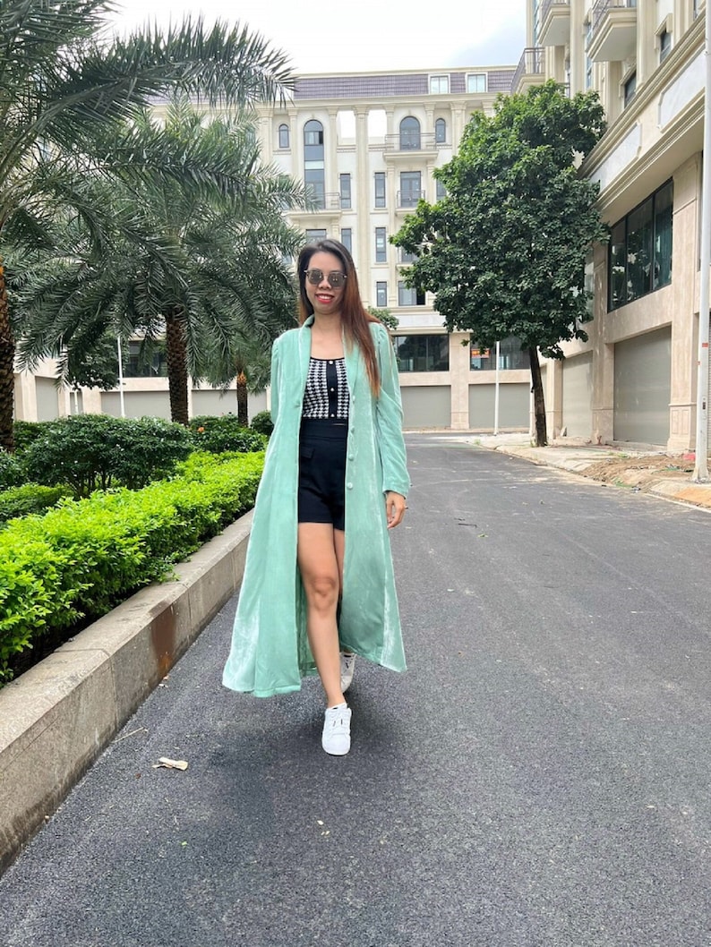 May include: A woman wearing a long, light green velvet duster coat over a black and white checkered top and black shorts. She is walking on a paved path with a building in the background.