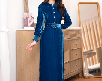 May include: A woman wearing a long, blue velvet dress with a white collar and a belt. The dress has a button-up front and long sleeves. She is standing in a room with a wooden floor and a dresser in the background.