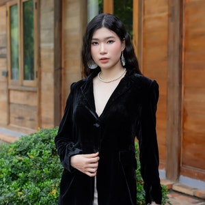 Pode incluir: Uma mulher vestindo um blazer de veludo preto com um colar de p&eacute;rolas brancas e uma pulseira de p&eacute;rolas brancas. Ela est&aacute; em p&eacute; na frente de um pr&eacute;dio de madeira com plantas verdes.