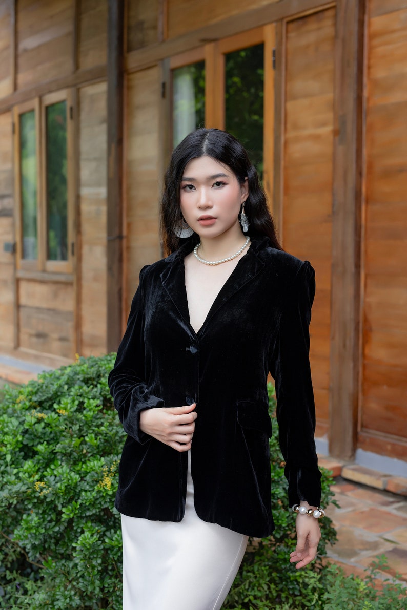 Pode incluir: Uma mulher vestindo um blazer de veludo preto com um colar de p&eacute;rolas brancas e uma pulseira de p&eacute;rolas brancas. Ela est&aacute; em p&eacute; na frente de um pr&eacute;dio de madeira com plantas verdes.