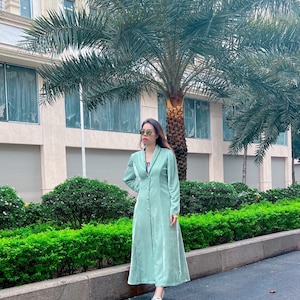 May include: A woman wearing a light green velvet dress with a button-up front and long sleeves. She is standing in front of a building with palm trees.