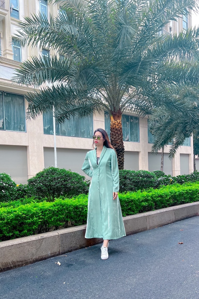 May include: A woman wearing a light green velvet dress with a button-up front and long sleeves. She is standing in front of a building with palm trees.