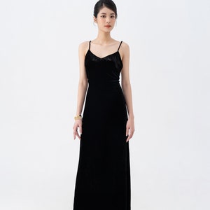 May include: A woman wearing a black velvet maxi dress with thin straps. She is standing on a white background and has her hair pulled back.