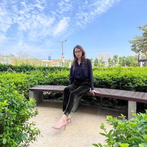 May include: A woman wearing a black blouse and dark green velvet pants sits on a wooden bench in a park. She is wearing glasses and has her hair pulled back. The bench is surrounded by green bushes and there is a blue sky in the background.