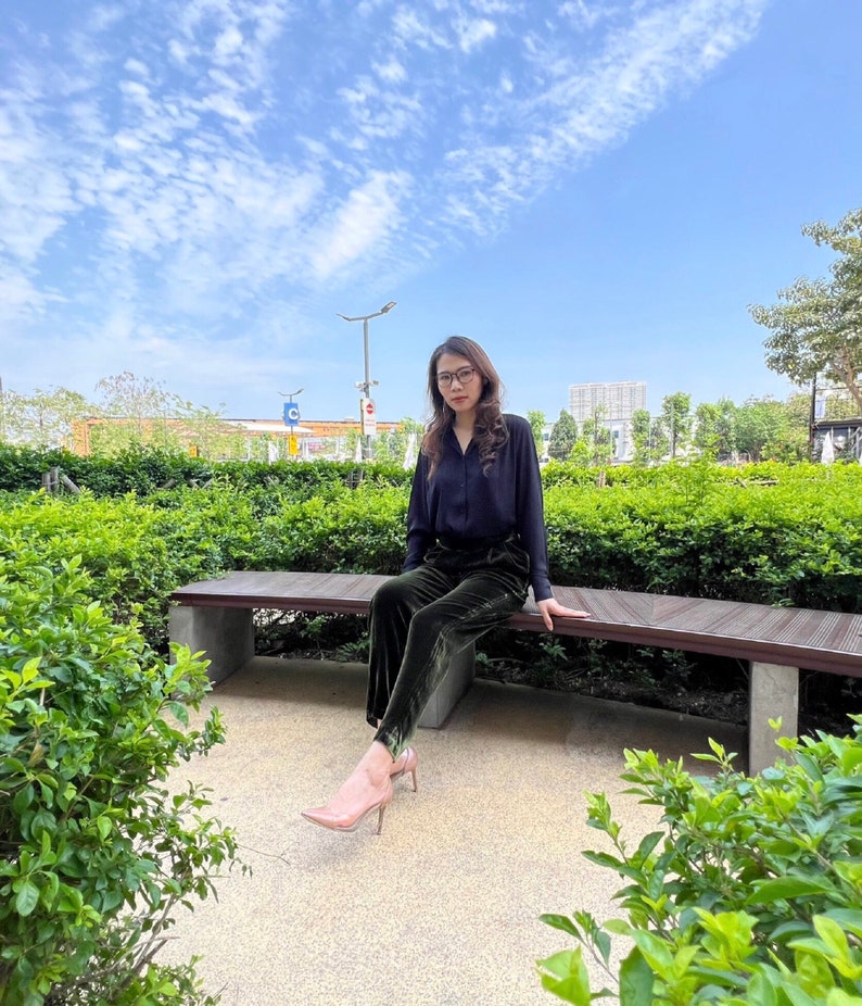 May include: A woman wearing a black blouse and dark green velvet pants sits on a wooden bench in a park. She is wearing glasses and has her hair pulled back. The bench is surrounded by green bushes and there is a blue sky in the background.