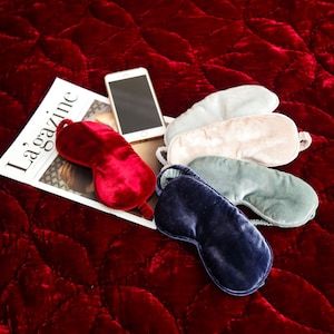 May include: Five different colored sleep masks made of velvet, laid out on a red quilted surface. The masks are in shades of red, pink, blue, gray, and white. A smartphone and a magazine titled "La Gazine" are also on the surface.
