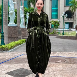 May include: A woman wearing a long, dark green velvet dress with a button-up front, a belt, and a hood. She is standing in front of a building with large windows.