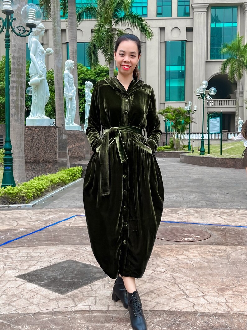 May include: A woman wearing a long, dark green velvet dress with a button-up front, a belt, and a hood. She is standing in front of a building with large windows.