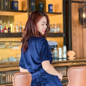 May include: A woman wearing a navy blue satin jumpsuit with a belt. She is standing in front of a bar with shelves behind her.