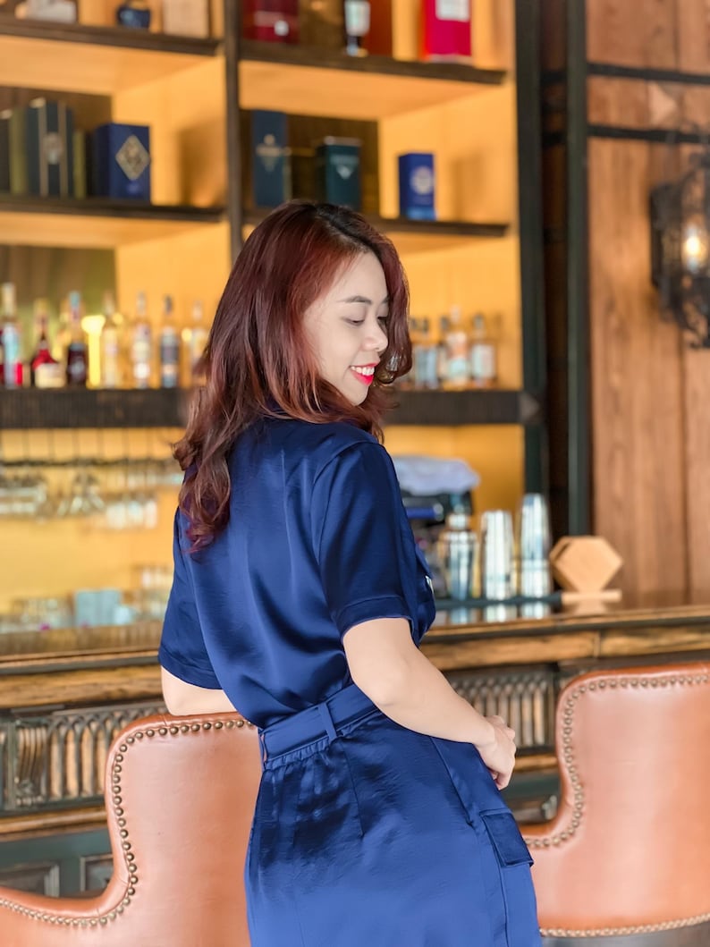 May include: A woman wearing a navy blue satin jumpsuit with a belt. She is standing in front of a bar with shelves behind her.