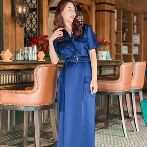 May include: A woman wearing a long, navy blue dress with a belt and pockets. She is standing in front of a bar with brown leather bar stools.