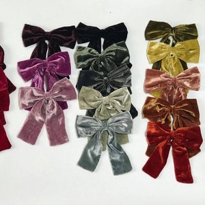May include: A collection of 15 velvet hair bows in various colors, including burgundy, pink, green, blue, and gold. The bows are arranged in rows on a white surface.