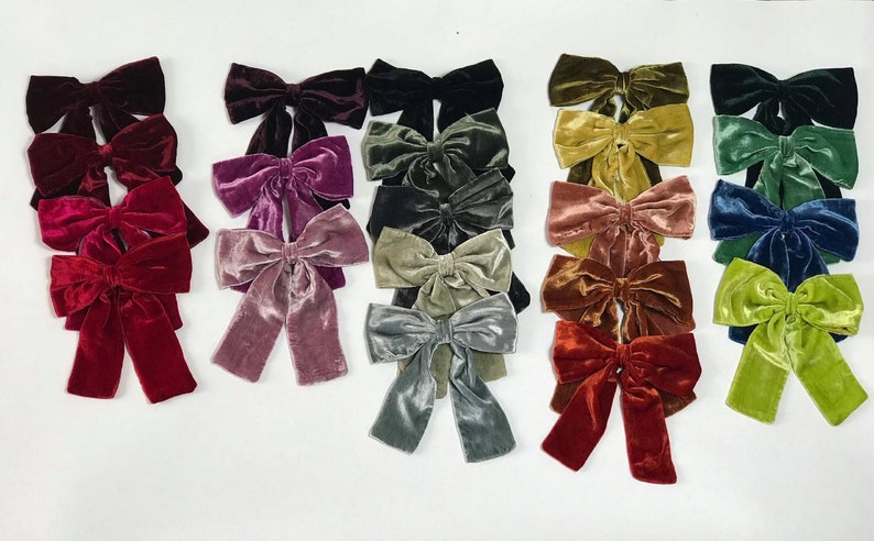 May include: A collection of 15 velvet hair bows in various colors, including burgundy, pink, green, blue, and gold. The bows are arranged in rows on a white surface.