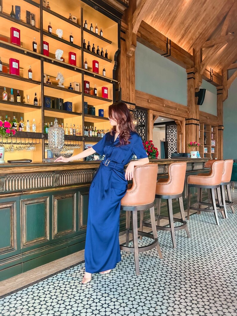 May include: A woman in a blue jumpsuit sits at a bar with a green and gold counter. The bar has a shelf with bottles of liquor behind it. The floor is tiled with a black and white geometric pattern.