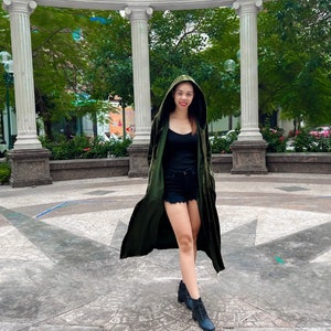 May include: A woman wearing a long, green velvet hooded jacket, a black tank top, and black denim shorts. She is standing in front of a white stone archway with green trees in the background.