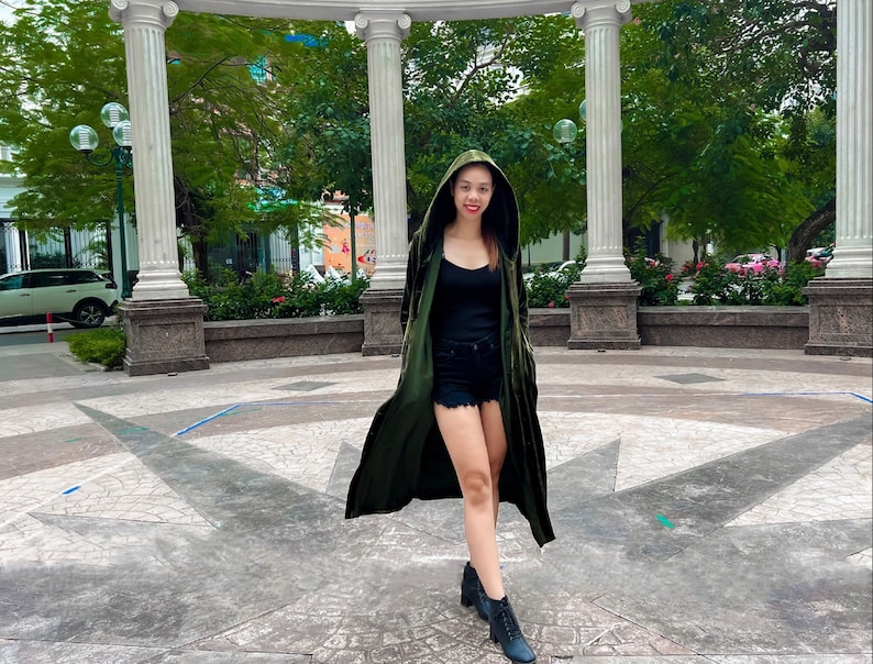 May include: A woman wearing a long, green velvet hooded jacket, a black tank top, and black denim shorts. She is standing in front of a white stone archway with green trees in the background.
