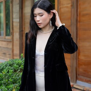 Pode incluir: Uma mulher vestindo um blazer de veludo preto sobre uma blusa de renda branca e uma saia branca. Ela est&aacute; usando brincos de p&eacute;rola e um colar de p&eacute;rola.