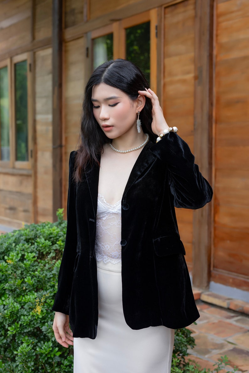 Pode incluir: Uma mulher vestindo um blazer de veludo preto sobre uma blusa de renda branca e uma saia branca. Ela est&aacute; usando brincos de p&eacute;rola e um colar de p&eacute;rola.