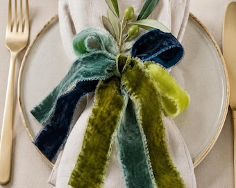 Puede incluir: Una servilleta de lino beige doblada sobre un plato de cerámica, atada con un conjunto de cintas de terciopelo deshilachadas en tonos azul marino, verde azulado y verde lima, decorada con una ramita de olivo y dos aceitunas verdes.