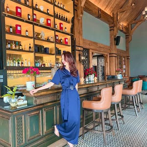 May include: A woman in a blue dress stands in a bar with a wooden ceiling and a green bar counter. The bar has a large shelf with bottles of liquor and a variety of glasses. The floor is tiled with a geometric pattern.