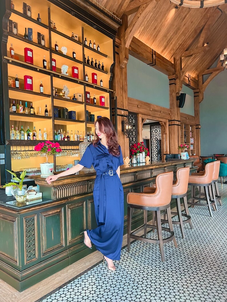 May include: A woman in a blue dress stands in a bar with a wooden ceiling and a green bar counter. The bar has a large shelf with bottles of liquor and a variety of glasses. The floor is tiled with a geometric pattern.