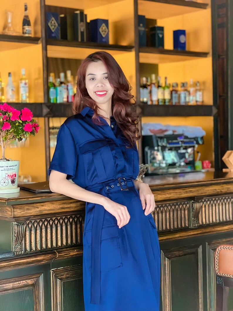 May include: A woman wearing a navy blue satin dress with a belt and pockets. She is standing behind a wooden counter with a green and gold design.