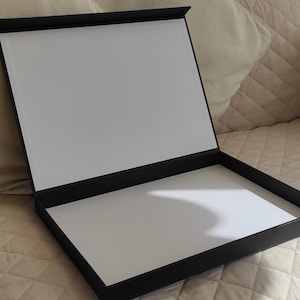 Może przedstawiać: Otwarta czarna skrzynka prezentacyjna z białym wnętrzem. Pudełko jest prostokątne z klapą na zawiasach, odsłaniającą białą powierzchnię wewnątrz. Pudełko spoczywa na beżowej pikowanej powierzchni.