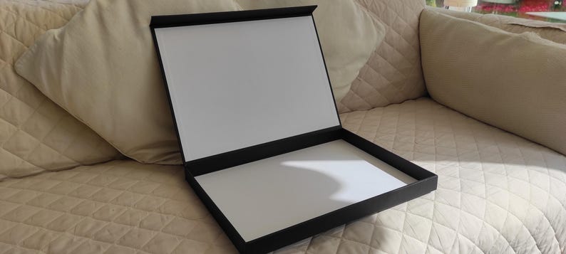 Peut inclure: Une bo&icirc;te de pr&eacute;sentation noire ouverte avec un int&eacute;rieur blanc. La bo&icirc;te est rectangulaire avec un couvercle &agrave; charni&egrave;re, r&eacute;v&eacute;lant une surface blanche &agrave; l'int&eacute;rieur. La bo&icirc;te repose sur une surface matelass&eacute;e beige.
