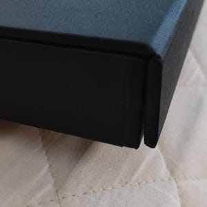 Peut inclure: Gros plan sur une bo&icirc;te noire &agrave; surface textur&eacute;e. La bo&icirc;te, avec un couvercle et une base, pr&eacute;sente une couture visible. Elle repose sur un tissu matelass&eacute; blanc cass&eacute;.